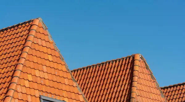Pomarańczowy dach wielospadowy z dachówki ceramicznej pod błękitnym niebem. Widoczny fragment okna dachowego.