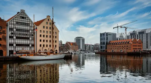 Widok na nabrzeże z zabytkowymi budynkami, żaglowcem i nowoczesną architekturą w tle.