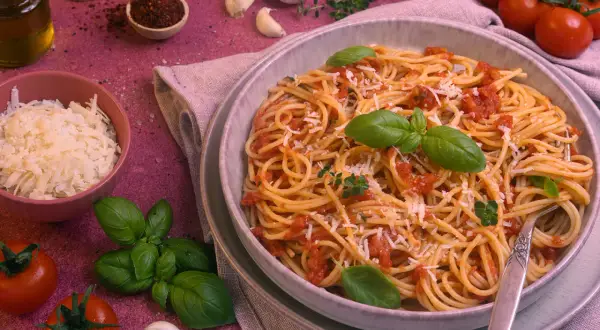 Ein Teller Spaghetti Napoli mit Tomatensauce, Basilikum und Parmesan. Daneben frische Tomaten, Knoblauch und Kräuter.