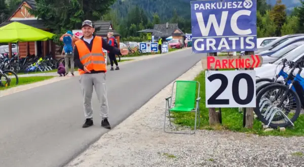Pracownik w kamizelce odblaskowej kieruje ruchem na parkingu siwa polana. W tle góry i budynki.