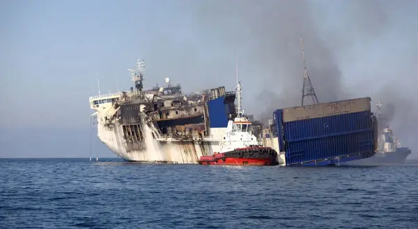 Beschädigtes Schiff mit Rauch, das von einem Schlepper im Meer gezogen wird.
