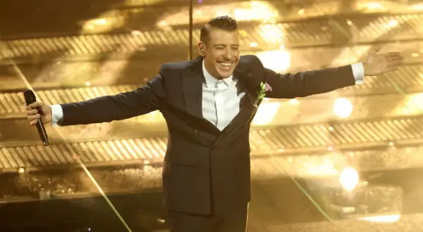 Un artista sorridente sul palco di Sanremo, con microfono in mano, pronto per i duetti.