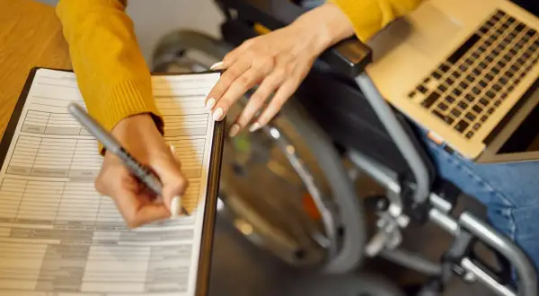 Kobieta na wózku inwalidzkim wypełnia dokumenty, korzystając z laptopa. To może być wniosek o 500 plus dla niepełnosprawnych.