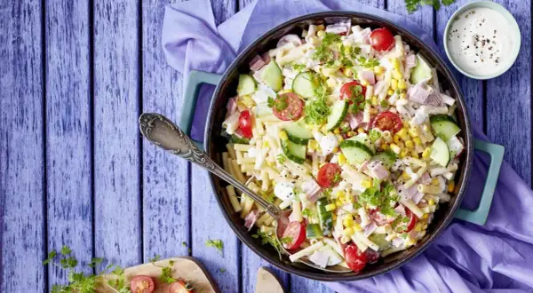 Ein köstlicher nudelsalat mit schinken, garniert mit Kirschtomaten, Gurkenscheiben und Mais. Perfekt für ein Sommerfest!
