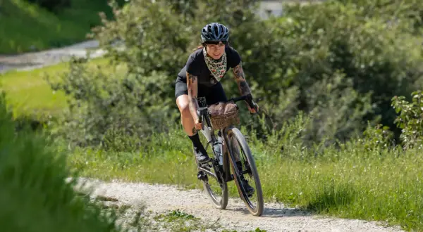 Kobieta z tatuażami na rowerze, ciesząca się kolarstwem gravelowym na piaszczystej drodze wśród zieleni.