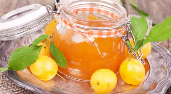 Ein Glas mit leuchtend orangefarbener, raffinierter Mirabellenmarmelade, umgeben von frischen Mirabellen und grünen Blättern auf einem silbernen Teller.