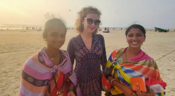 Trzy kobiety na plaży w Indiach. Jedna z nich trzyma pomarańczowy notes, może to notatnik do zapisywania cen rupii, czyli waluty w Indiach.