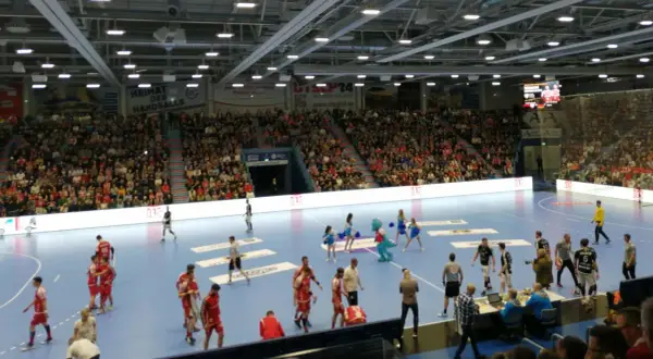 Handballspiel in der Gummersbach Schwalbe Arena. Zuschauer füllen die Ränge, Spieler auf dem Feld, eine Maskottchen-Figur und Cheerleader sind zu sehen.