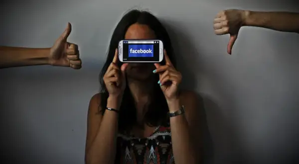Una persona oculta su rostro tras un móvil con Facebook. A los lados, pulgares arriba y abajo, reflejando el impacto de las redes sociales y la autoestima.