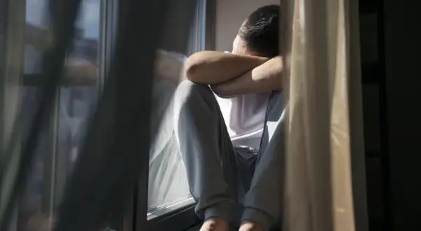 Mężczyzna siedzi przy oknie, obejmując się ramionami. Światło słoneczne pada na jego twarz, podkreślając jego lęk przed wyjściem z domu.