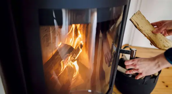 Ein Kaminofen mit brennendem Holz. Eine Hand hält ein Holzscheit, bereit zum Nachlegen. Bald muss der alte Kaminofen entsorgt werden.