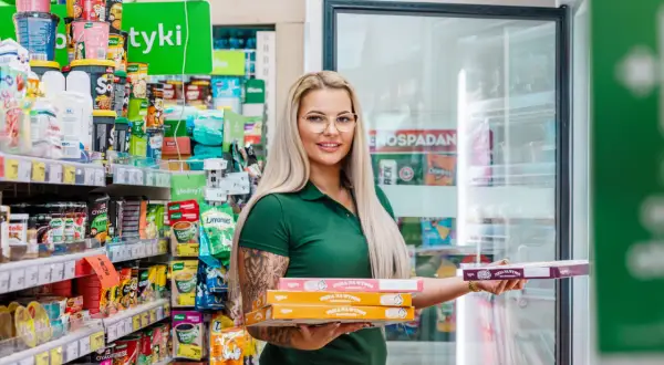 Kobieta w zielonej koszulce trzyma pudełka z pizzą na wynos w sklepie spożywczym.