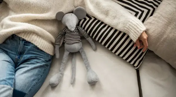 Graues Stofftier-Mäuschen mit gestreiftem Oberteil liegt neben Person in Strickpullover und Jeans auf Sofa.