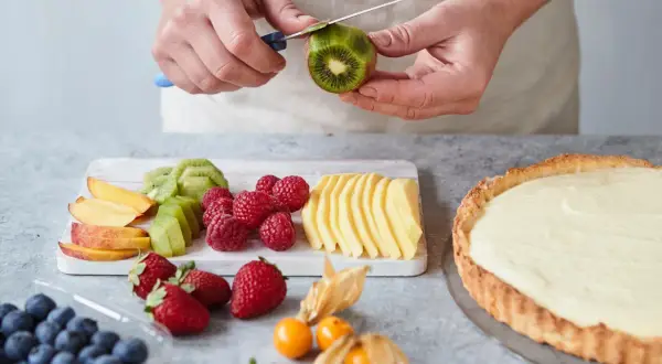 Hände schälen Kiwi für ein fruchtiges Tarte Rezept. Daneben liegen Beeren, Pfirsichspalten und Apfelscheiben.
