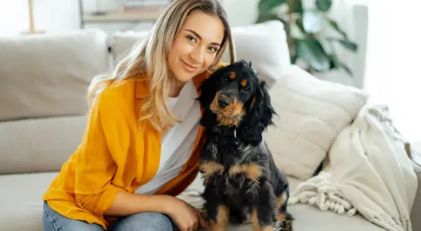 Uśmiechnięta blondynka i jej piesek siedzą na kanapie. Piesek patrzy w obiektyw, jakby chciał powiedzieć: "dlaczego pies chodzi za mną krok w krok?".