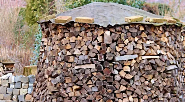 Ein großer Stapel Brennholz, der mit einer Plane und Ziegeln abgedeckt ist. Die Holzscheite sind ordentlich aufgeschichtet, bereit für den Winter. Die Baustahlmatte dient als stabile Unterlage.