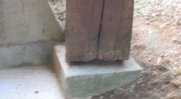 Un poteau bois pourri à la base repose sur un socle en béton. La dégradation du bois est visible.