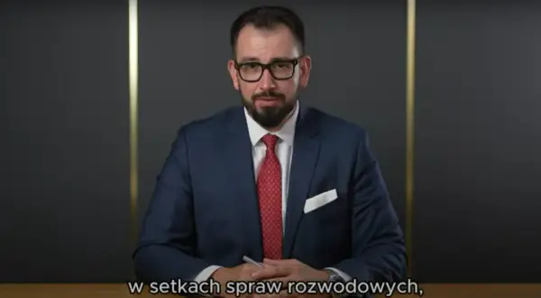 Mężczyzna w garniturze i okularach siedzi przy biurku, trzymając długopis.