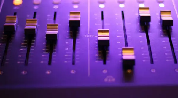 Panel miksera dźwiękowego z faderami i podświetlanymi przyciskami w fioletowym świetle.