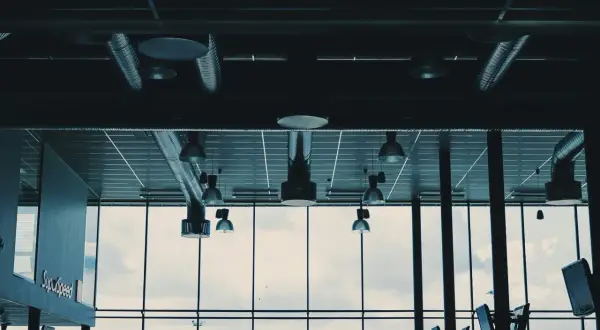 Ventilationsrör och lampor i taket, med en stor fönstervägg som visar en molnig himmel och en grönskande kulle. Kanske är det en plats där man kontrollerar radon nivåer.