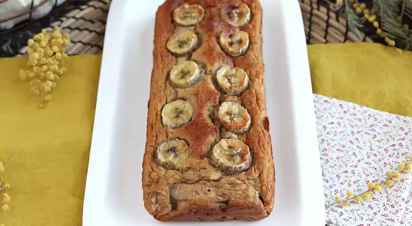 Delizioso banana bread light, decorato con fette di banana, su un piatto bianco, con fiori di mimosa sullo sfondo.