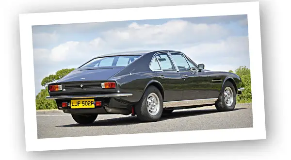 Klasyczny Aston Martin Lagonda w ciemnym kolorze, widziany z tyłu, na tle błękitnego nieba z chmurami.