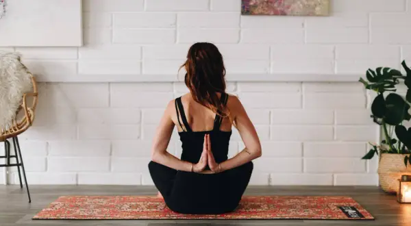 Femme en tenue noire pratiquant le hatha yoga, mains jointes derrière le dos, assise sur un tapis orné.