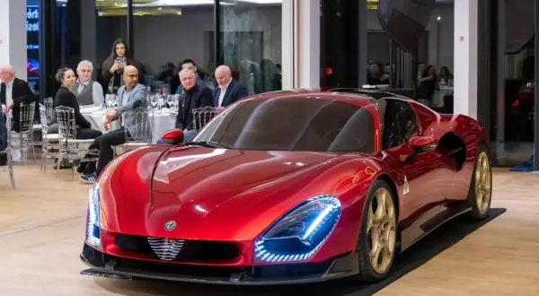 Lśniący, czerwony Alfa Romeo 33 z złotymi felgami prezentuje się na tle sali bankietowej, gdzie goście przyglądają się mu z zaciekawieniem.