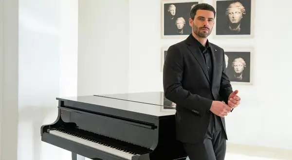 Uomo in elegante outfit teatro, con completo nero, accanto a un pianoforte a coda.