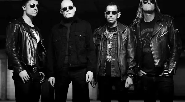 Czterech mężczyzn w skórzanych kurtkach, w tym jeden z napisem "Sisters of Mercy" na koszulce, pozuje do zdjęcia.