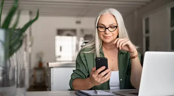 Frau mit Brille und Handy am Tisch, plant das Haus verkaufen und in Mietwohnung ziehen.