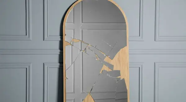 A tall, arched mirror with a wooden frame is shattered, with many glass shards reflecting a grey, paneled wall.