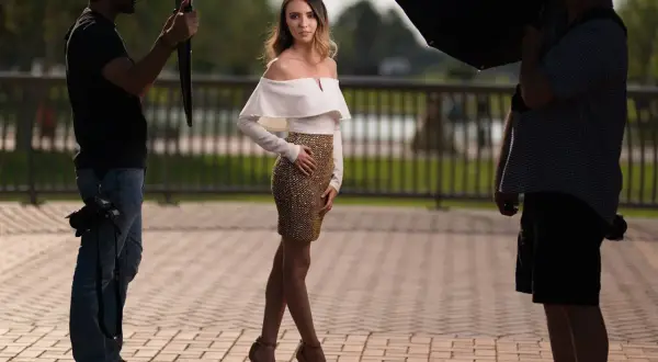 Modelka w złotej spódnicy i białej bluzce pozuje do sesji. Fotografowie z lampą i parasolką dbają o idealne światło. TTL co to za magia?