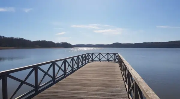 Holzsteg am Schmachter See lädt zum Spaziergang auf dem Rundweg ein. Sonnenschein glitzert auf dem Wasser, bewaldete Ufer im Hintergrund.