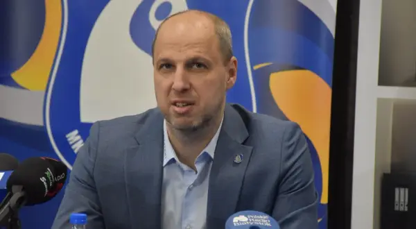 Dominik Kwapisiewicz w garniturze przed mikrofonami, na tle niebiesko-żółtego logo.