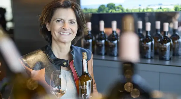 Frau mit Weinflasche und Glas, im Hintergrund Flaschenreihe und Landschaft.