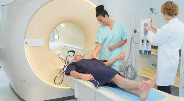 Pacjent leży na łóżku przed aparatem MRI. Dwie pielęgniarki przygotowują go do badania. To właśnie mri co to za badanie.