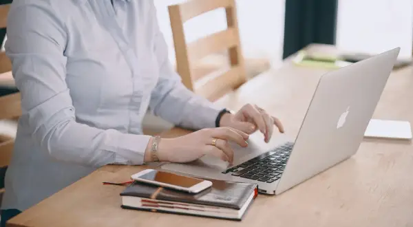 Kobieta w jasnej koszuli pracuje na laptopie, obok leży telefon i notatnik. Dba o swoje ubezpieczenie chorobowe.