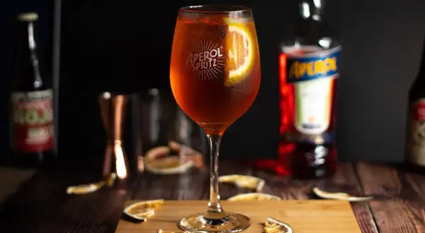 Orzeźwiający Aperol Spritz w kieliszku, ozdobiony plasterkiem pomarańczy. Obok butelka Aperol i suszone pomarańcze.