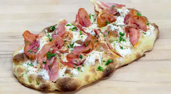 Une délicieuse pinsa recette garnie de burrata crémeuse, de fines tranches de prosciutto et de persil frais, le tout sur une pâte dorée.