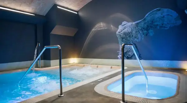 Piscinas de hidromasaje iluminadas con luz azul, con barandillas metálicas. Un mural de una escultura antigua adorna la pared, sugiriendo el mejor spa de Valencia.