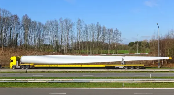 Żółta ciężarówka transportuje ogromne łopaty wiatraka. Ten pojazd nienormatywny pokonuje drogę, by dostarczyć element do budowy farmy wiatrowej.
