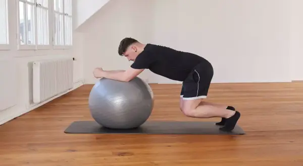Un homme effectue un exercice de gainage avec un ballon de gym, en appui sur ses avant-bras et ses genoux.