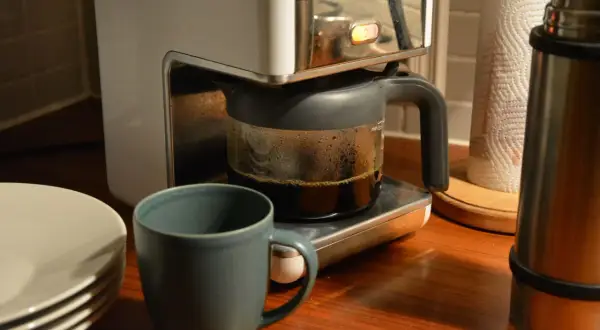 En kaffebryggare med en glaskanna full med färskt kaffe. Bredvid står en mugg och en stapel tallrikar. Man kan brygga presskaffe i kaffebryggare för en god start på dagen.