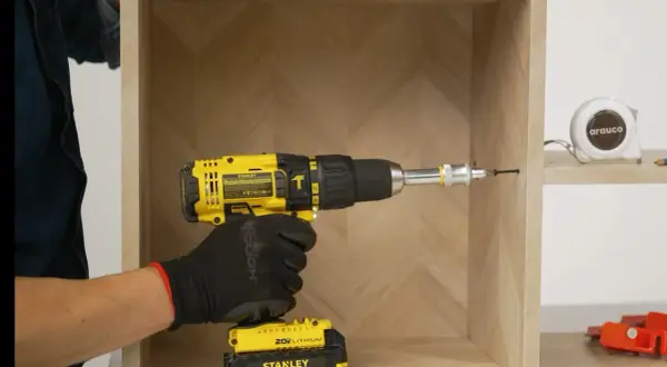 Mano con guante negro usa taladro amarillo para unir dos muebles. Cinta métrica y herramientas al fondo.