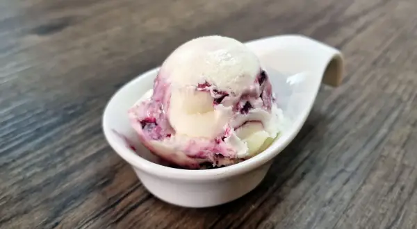 Cremiges Cheesecake Eis mit fruchtigen Beerenwirbeln in einer weißen Schale auf Holztisch.