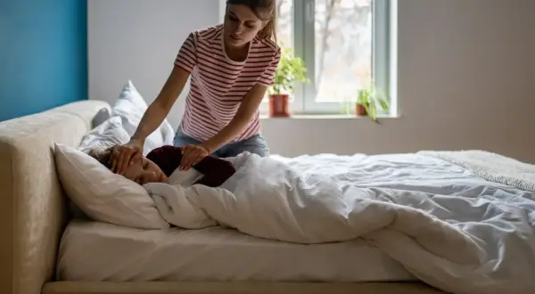 Kobieta sprawdza czoło śpiącego dziecka, które leży w łóżku pod kołdrą. Wygląda na to, że dziecko ma przeziębienie.