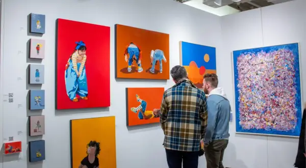 Two men examine colorful paintings at an art fair, perhaps contemplating their next acquisition as an art collector.