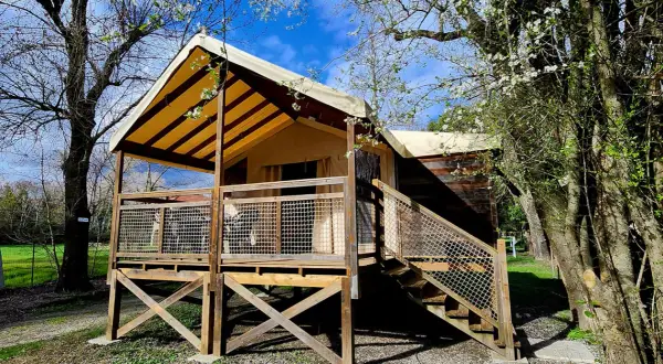 Un lodge de camping éco-responsable avec une tente surélevée et une terrasse en bois, entouré d'arbres fleuris sous un ciel bleu.