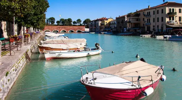 Barche ormeggiate nel canale, un assaggio di cosa vedere a Peschiera del Garda. Edifici colorati e un ponte ad arcate sullo sfondo.